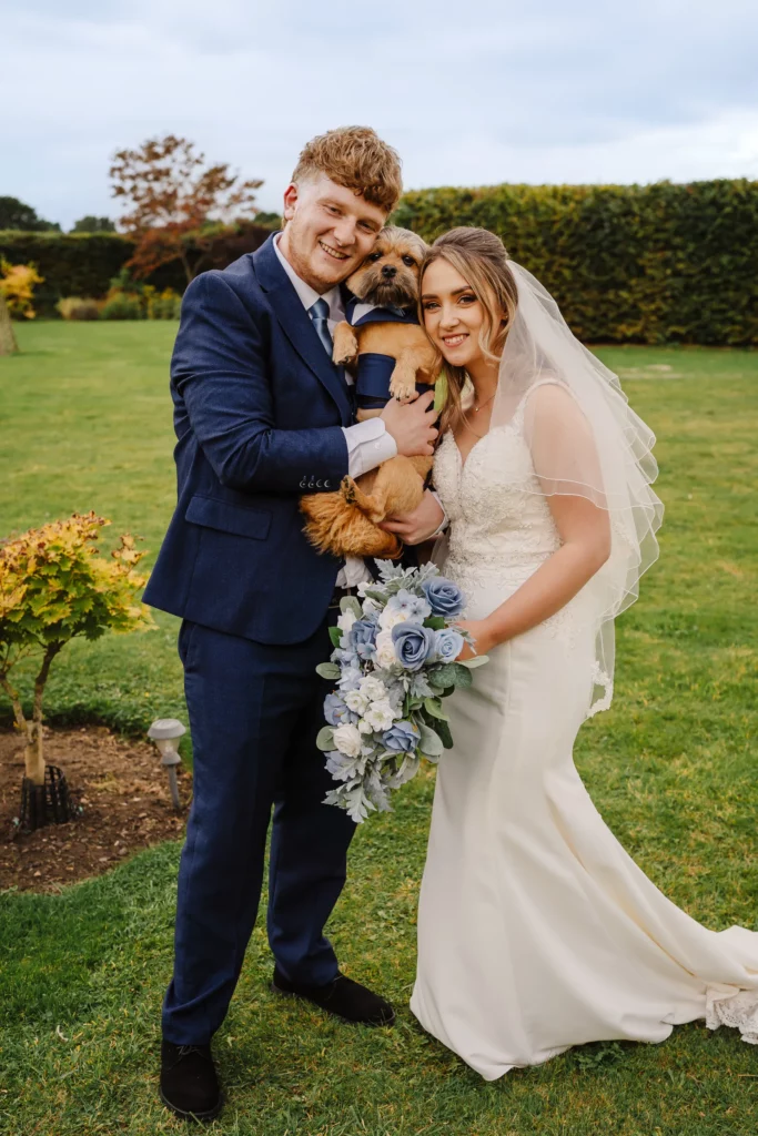 A bride in a white gown and veil stands beside a groom in a navy suit. They smile while holding a small brown dog dressed in a suit. The bride holds a bouquet of blue and white flowers. They are outdoors on a grassy lawn. Bethany Maeve Photography