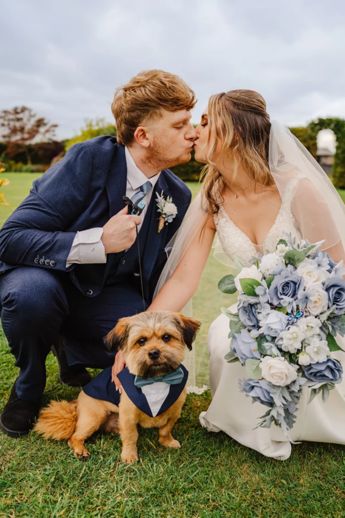 A bride and groom kneel on grass, dressed in wedding attire, sharing a kiss. In front of them sits a small brown dog in a blue tuxedo, and the bride holds a bouquet of white and blue flowers. Bethany Maeve Photography