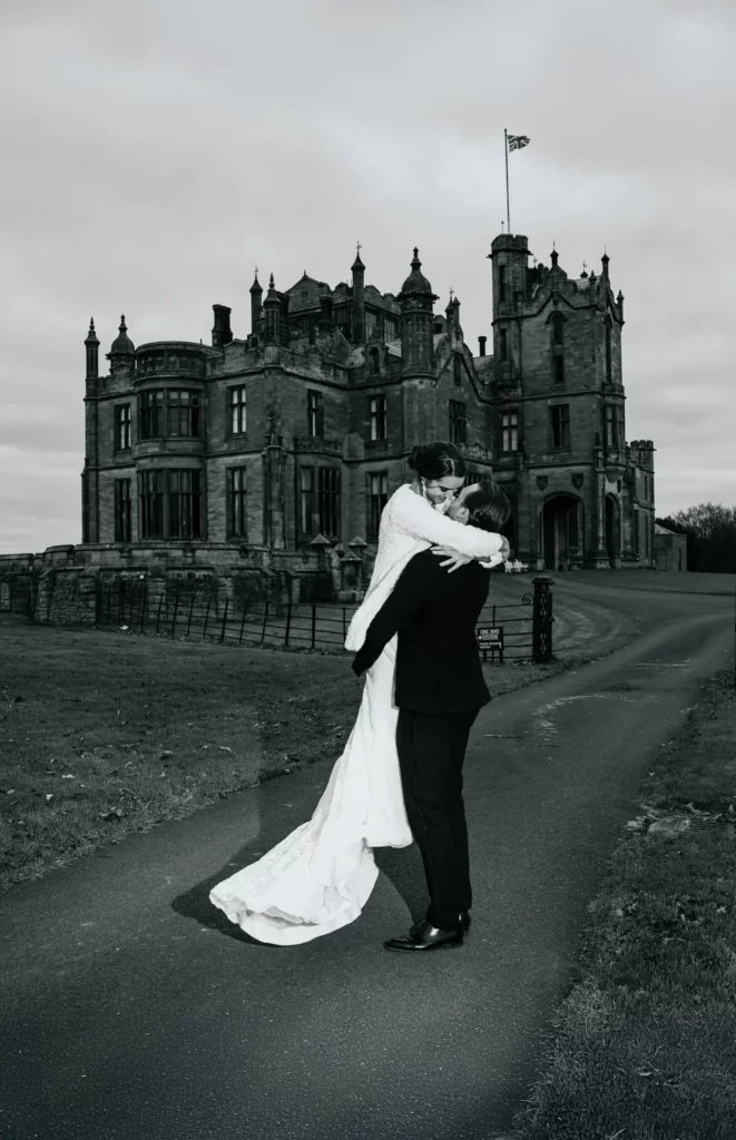 A bride and groom embrace joyfully on a path in front of a grand, historic castle. The photo is black and white, highlighting the elegance and romance of their wedding day. Bethany Maeve Photography