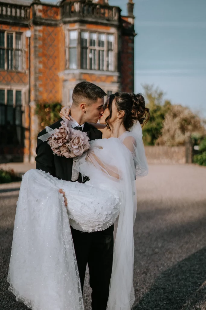 A groom in a black suit carries his bride in a white wedding dress and veil outside a historic building. They smile and gaze at each other lovingly, with the bride holding a bouquet of flowers. Bethany Maeve Photography