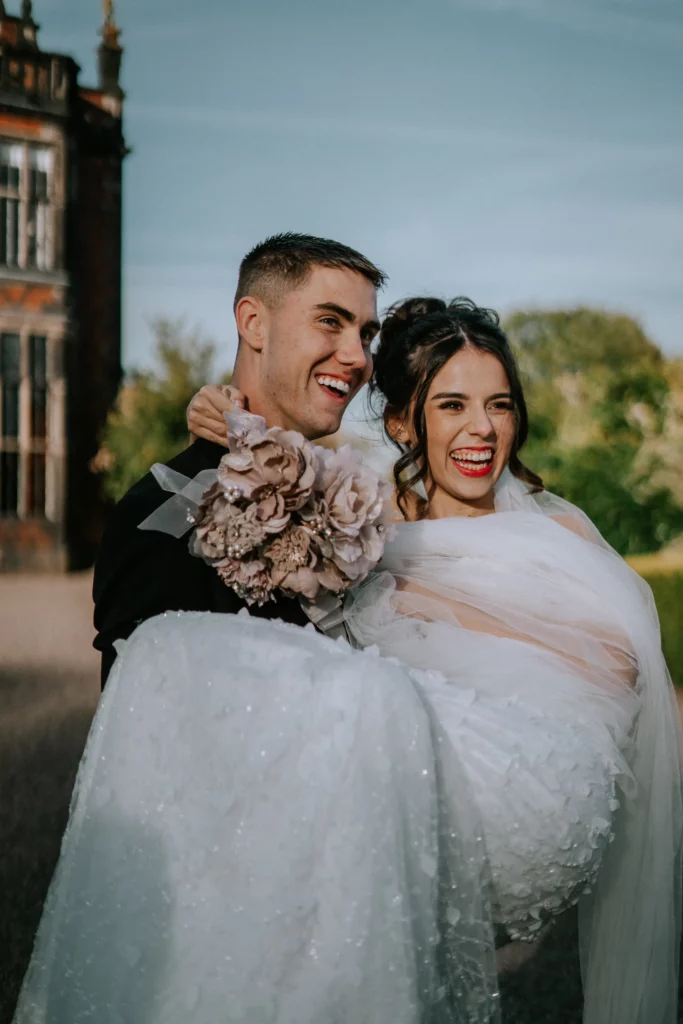 A groom in a black suit joyfully carries his smiling bride in a white wedding dress and veil outdoors. The bride holds a bouquet of flowers, and they both look happy against a green, sunlit background. Bethany Maeve Photography
