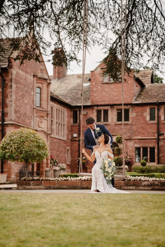 A bride in a white dress sits on a swing holding a bouquet whilst a groom in a blue suit stands behind her, in front of a large historic brick building with arched windows and flowers. Bethany Maeve Photography