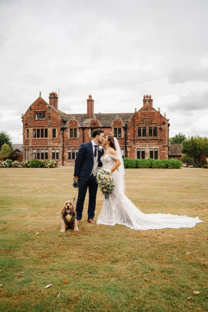 A bride and groom stand close together, smiling with their dog on a lead in front of a large, historic brick mansion on a cloudy day. The bride holds a bouquet and wears a long white dress and veil. Bethany Maeve Photography
