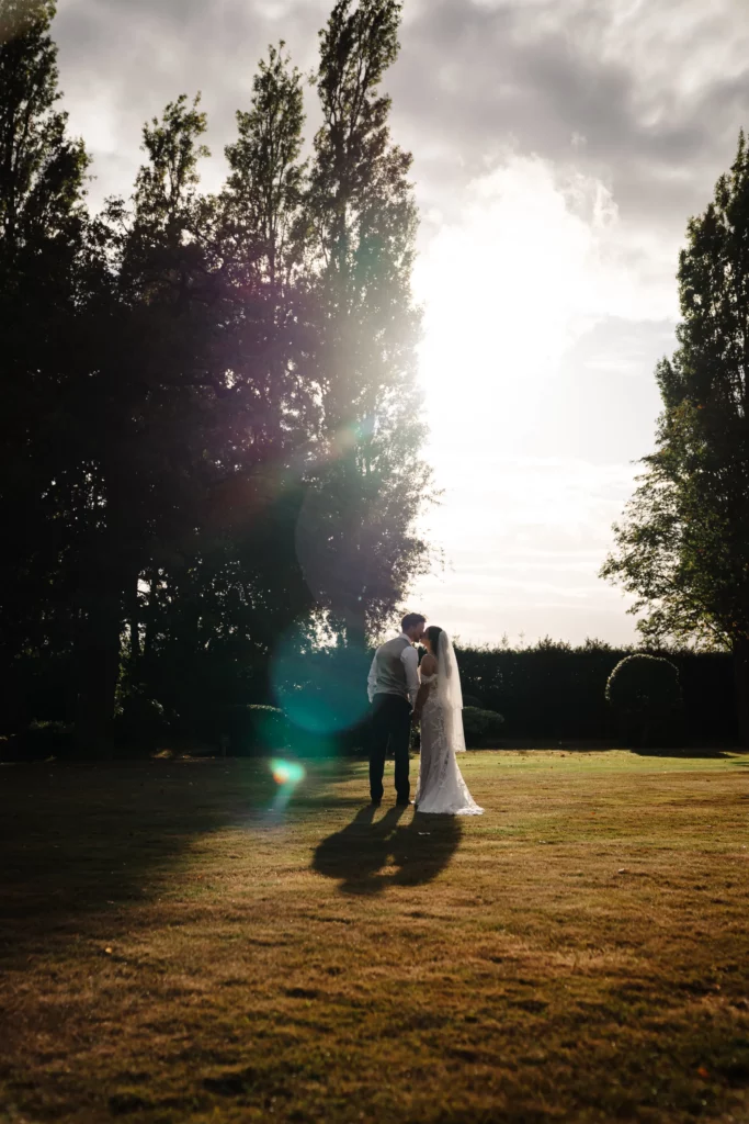 A bride and groom stand close together, kissing in the sunlight on a grassy lawn, with tall trees and a dramatic, cloudy sky in the background. Bethany Maeve Photography
