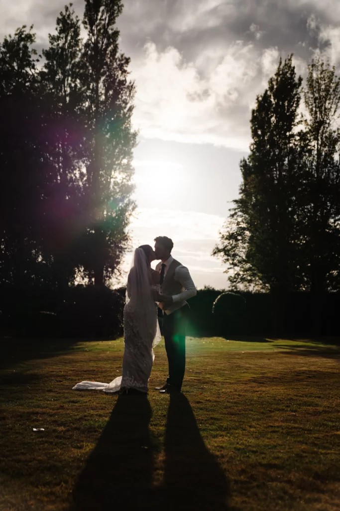 A bride and groom stand close together outdoors, silhouetted against the setting sun, with tall trees framing them and sunlight creating a soft glow around their figures. Bethany Maeve Photography