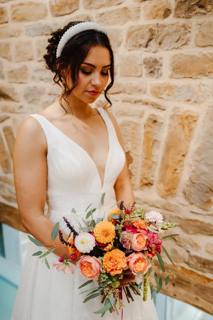 A bride in a white dress with a deep neckline holds a colourful bouquet of flowers, standing against a rustic stone wall and looking down thoughtfully. Bethany Maeve Photography