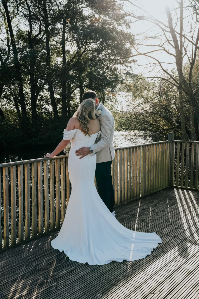 A bride in a white off-shoulder gown and a groom in a light suit embrace on a wooden deck, facing a sunlit lake surrounded by trees. Sunlight filters through the branches, casting a romantic glow. Bethany Maeve Photography