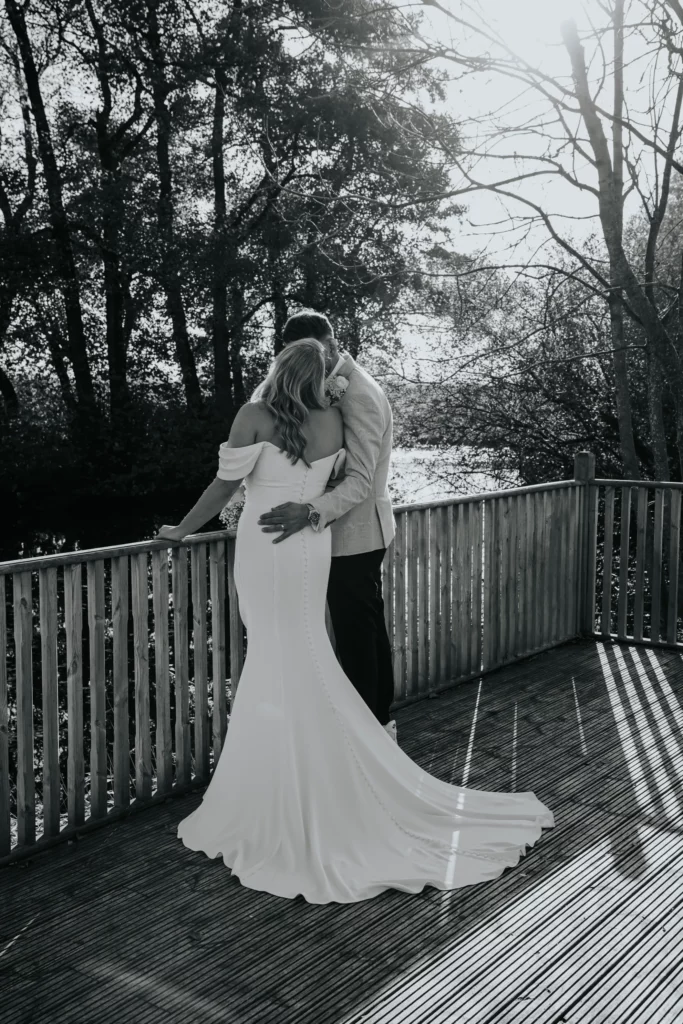 A couple stands on a wooden terrace, embracing closely. The woman wears a long, off-the-shoulder white dress, and the man wears a light suit. Sunlight streams through trees in the background, creating a romantic atmosphere. Bethany Maeve Photography