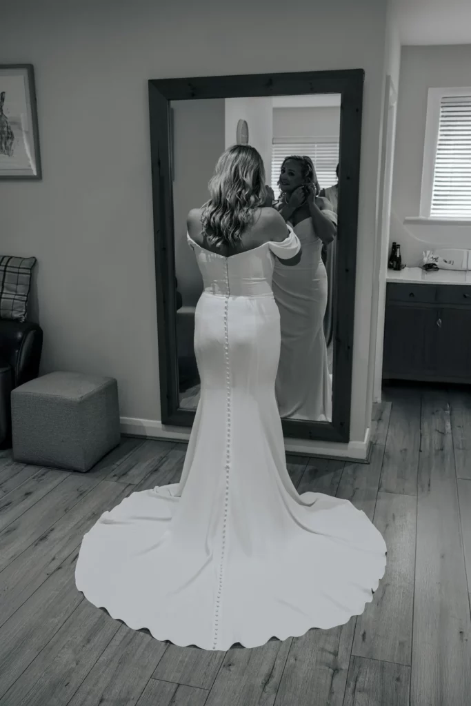 A woman in a white, off-the-shoulder wedding dress stands facing a large mirror, adjusting her earring. The back of her dress features a row of buttons and a long train. The room has wooden floors and simple decor. Bethany Maeve Photography