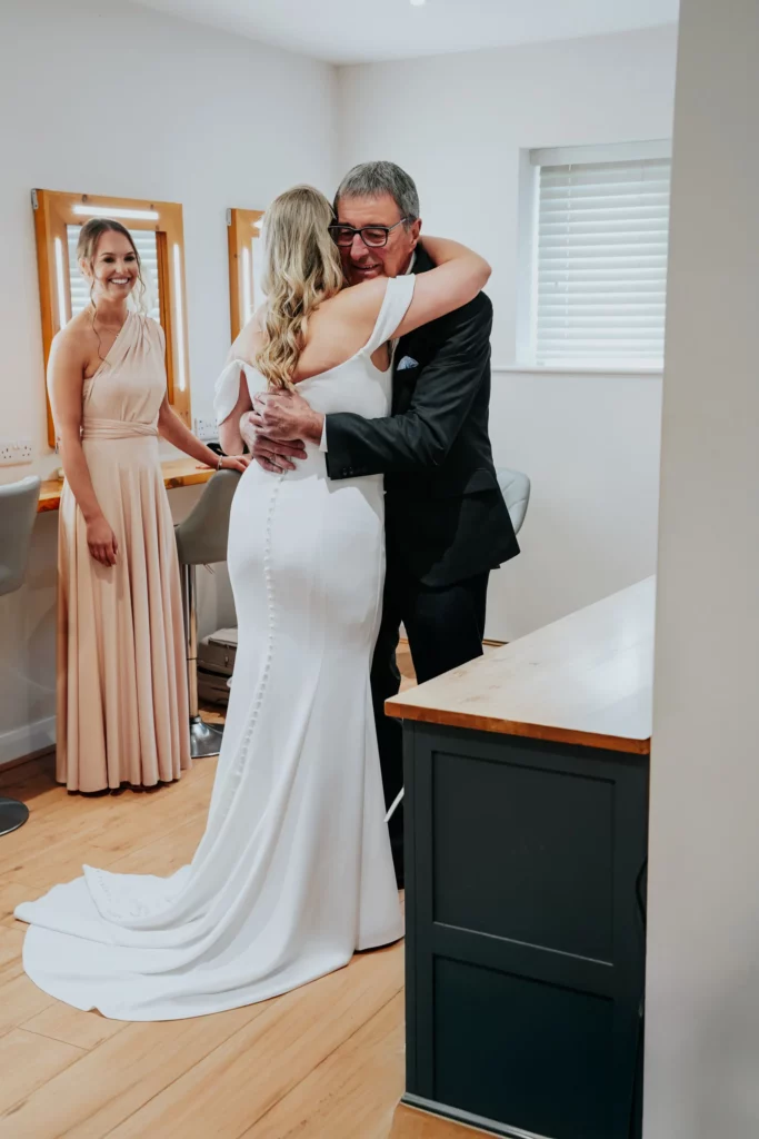 A bride in a white dress hugs an older man in a suit, both smiling. A bridesmaid in a light pink dress stands nearby, watching and smiling in a bright room with mirrors and wooden floors. Bethany Maeve Photography