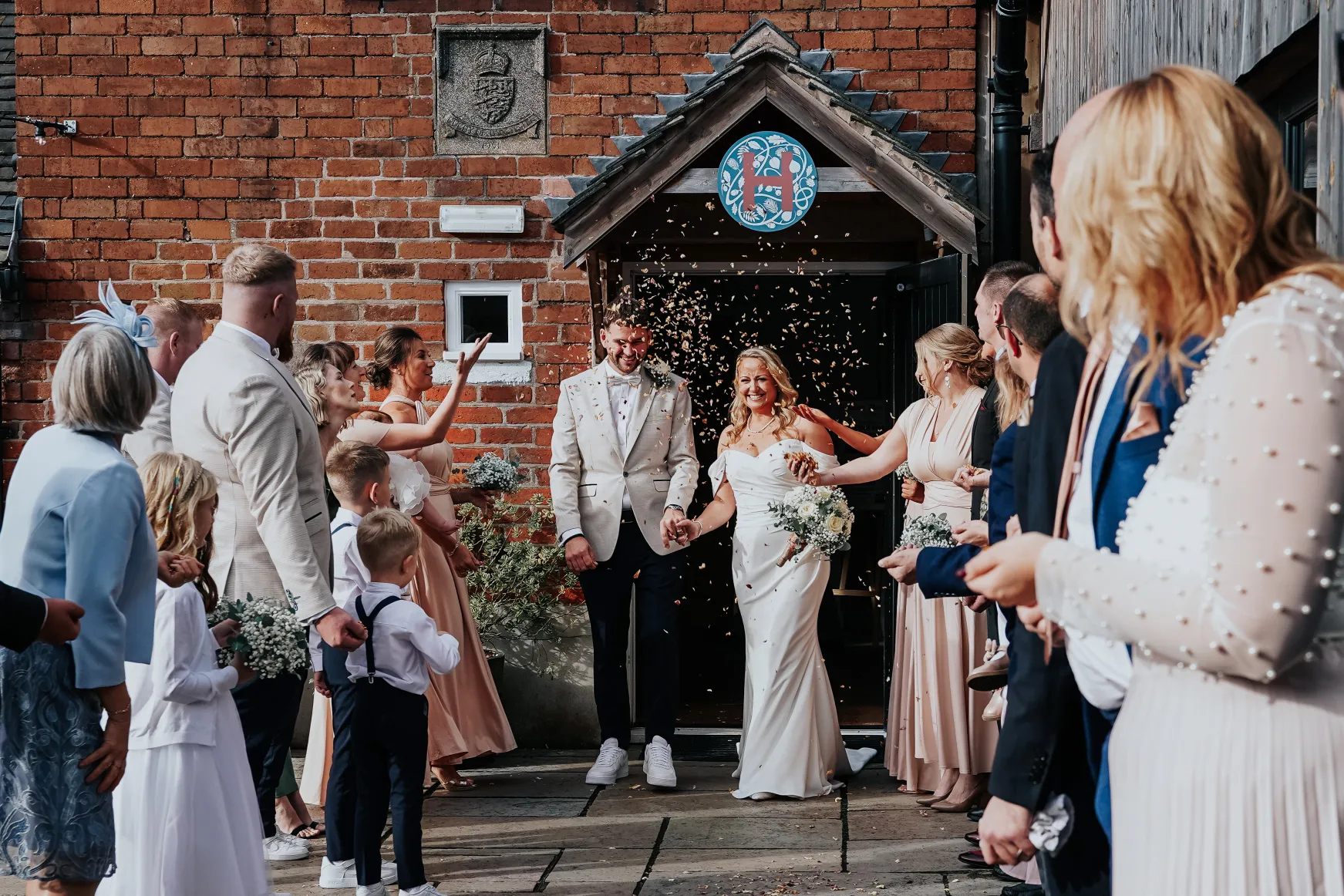 A newlywed couple smiles and walks hand in hand outside, surrounded by guests who are throwing confetti. The group celebrates joyfully near a rustic brick building with wooden features. Bethany Maeve Photography