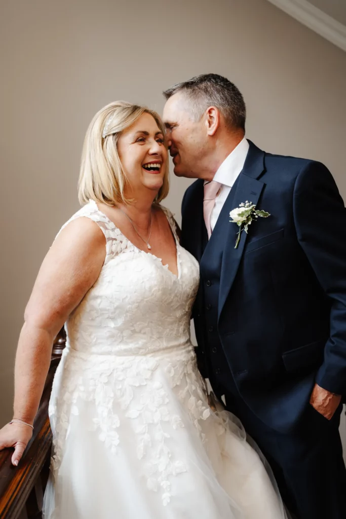 A smiling bride in a white lace wedding dress stands beside a groom in a dark suit with a buttonhole. He leans in close, appearing to whisper in her ear, making her laugh. Bethany Maeve Photography