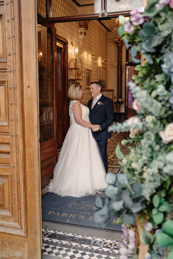 A bride and groom hold hands and smile at each other inside an elegant building, framed by a wooden door and a floral arrangement. The bride wears a white dress and the groom is in a dark suit. Bethany Maeve Photography