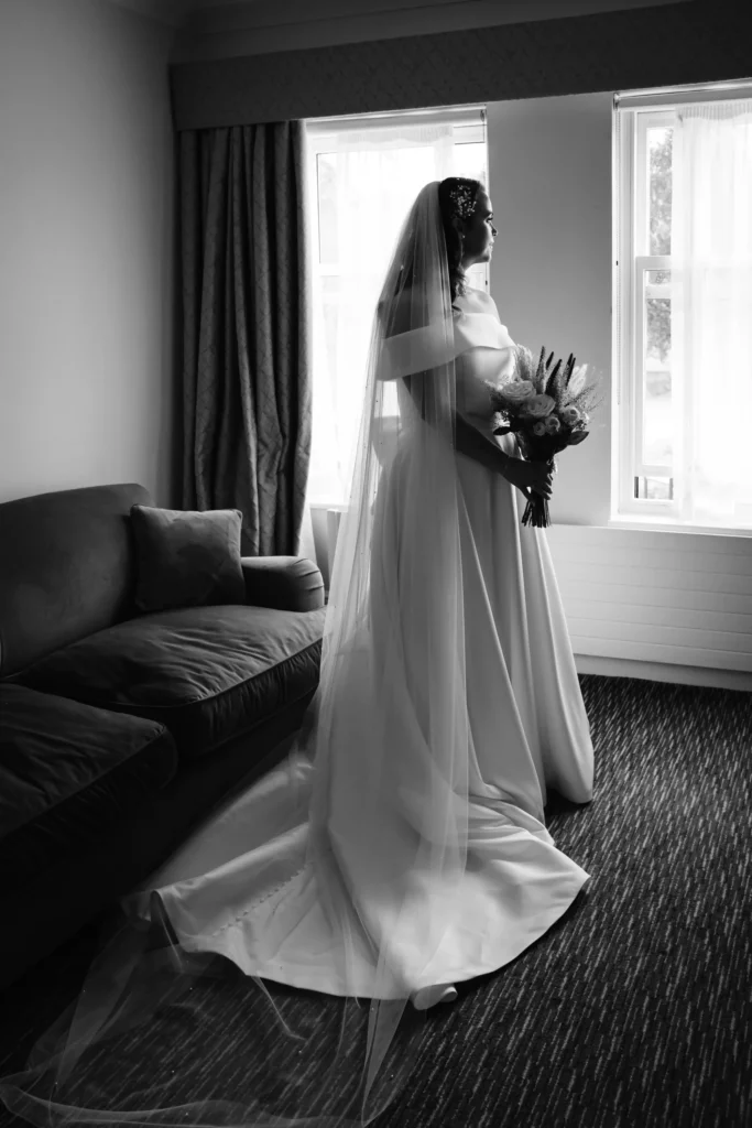 A bride in a long, elegant gown and veil stands by a window holding a bouquet, looking outside. Sunlight filters in, highlighting her silhouette in a softly lit room with a sofa and curtains. Black and white photo. Bethany Maeve Photography