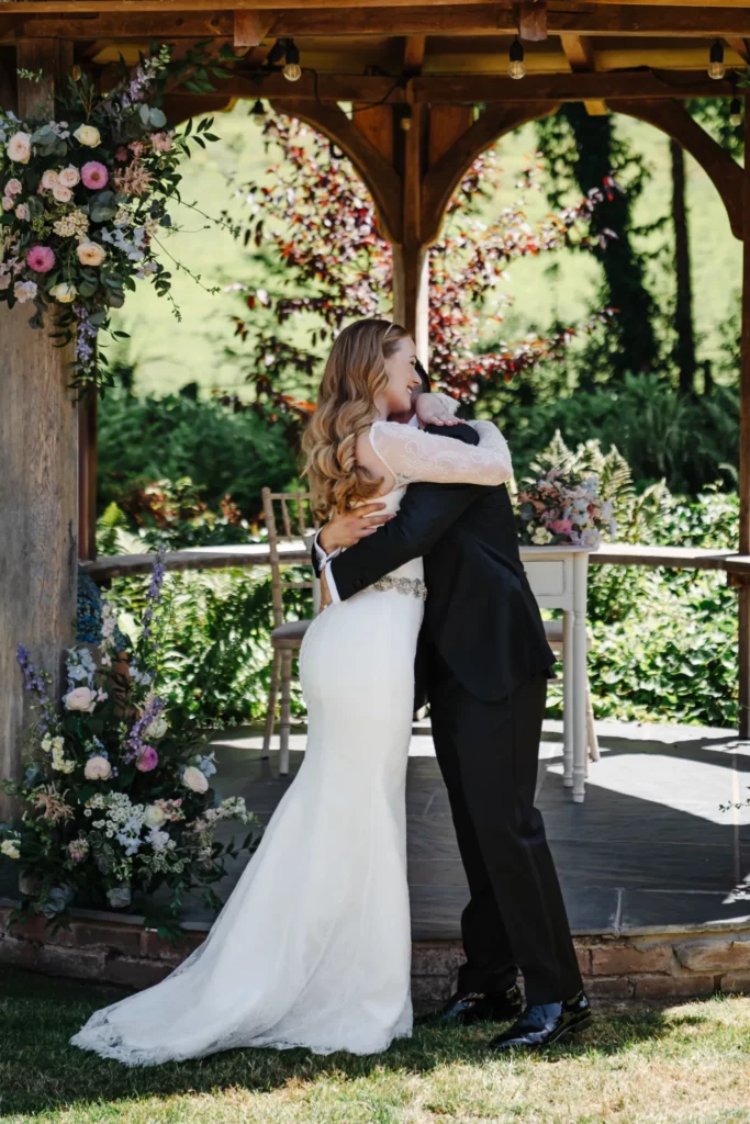 A bride in a white gown and a groom in a black suit embrace under a wooden gazebo decorated with flowers, surrounded by lush greenery and vibrant plants on a sunny day. Bethany Maeve Photography