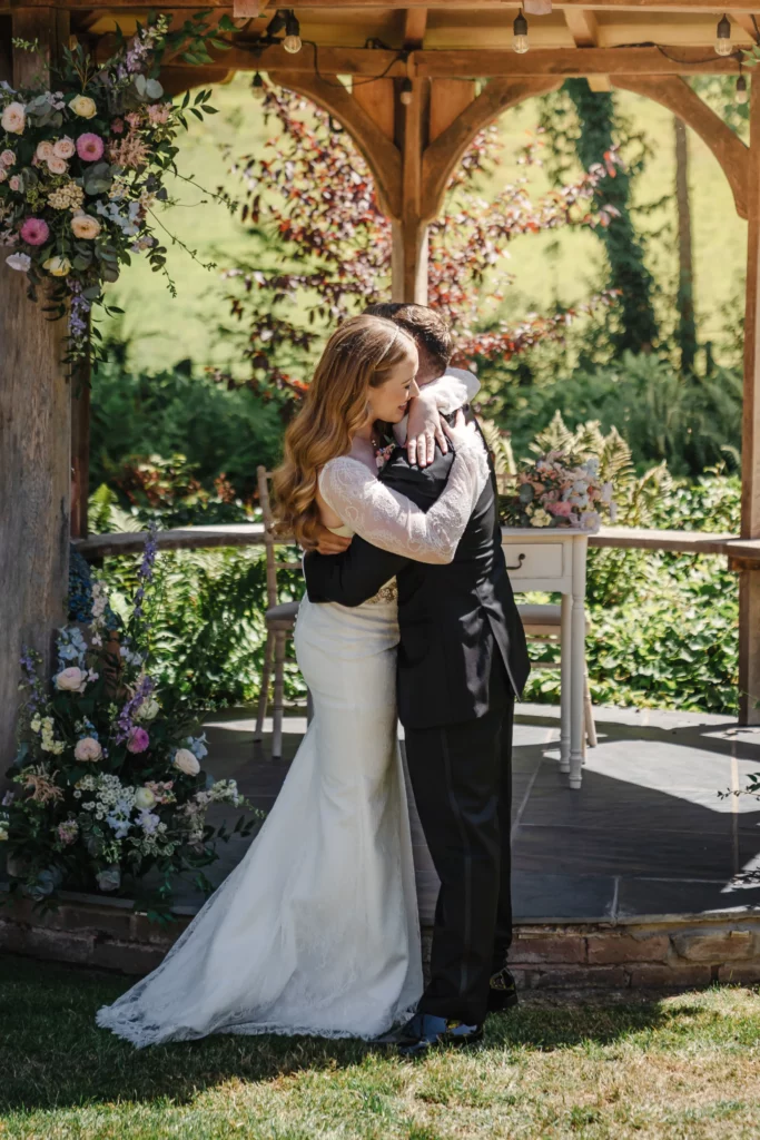 A bride in a white dress and a groom in a black suit embrace under a wooden gazebo adorned with floral arrangements, surrounded by greenery and sunlight. Bethany Maeve Photography