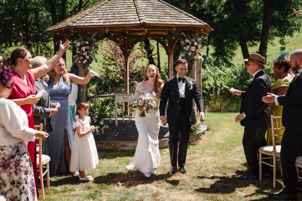 A bride and groom walk hand-in-hand down the aisle, smiling, as guests cheer and throw confetti. They are outdoors in front of a flower-decorated gazebo, surrounded by family and friends in celebratory attire. Bethany Maeve Photography