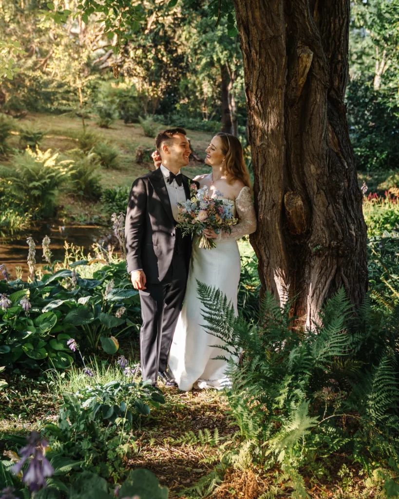A bride and groom stand together in a sunlit garden, smiling at each other by a large tree. The bride holds a bouquet of flowers, and both are dressed in formal wedding attire. Lush greenery surrounds them. Bethany Maeve Photography