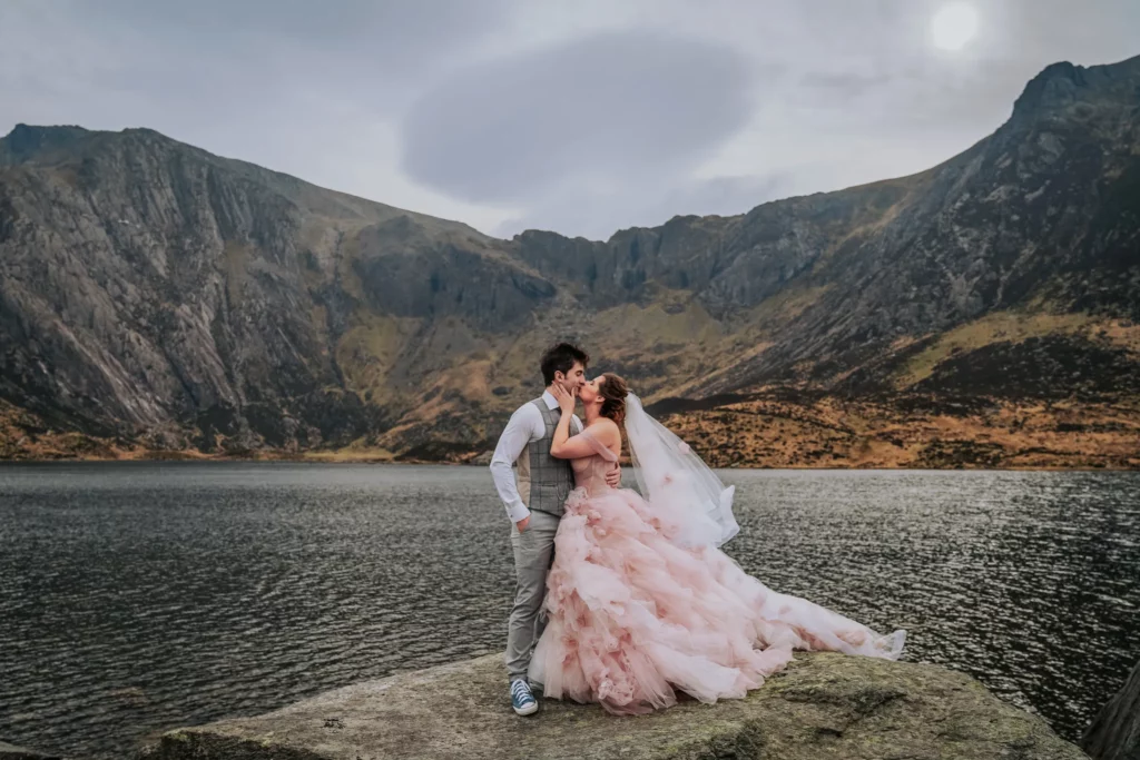 A couple in wedding attire kisses on a rock by a lake, surrounded by mountains. The bride wears a flowing pink dress and veil, the groom a grey waistcoat and trousers. The scene is dramatic and romantic under a cloudy sky. Bethany Maeve Photography