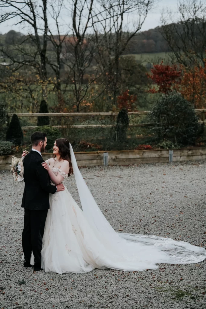 A bride in a long white dress and veil stands outdoors with a groom in a black suit. They smile at each other, holding hands, with trees and autumn foliage in the background. Bethany Maeve Photography