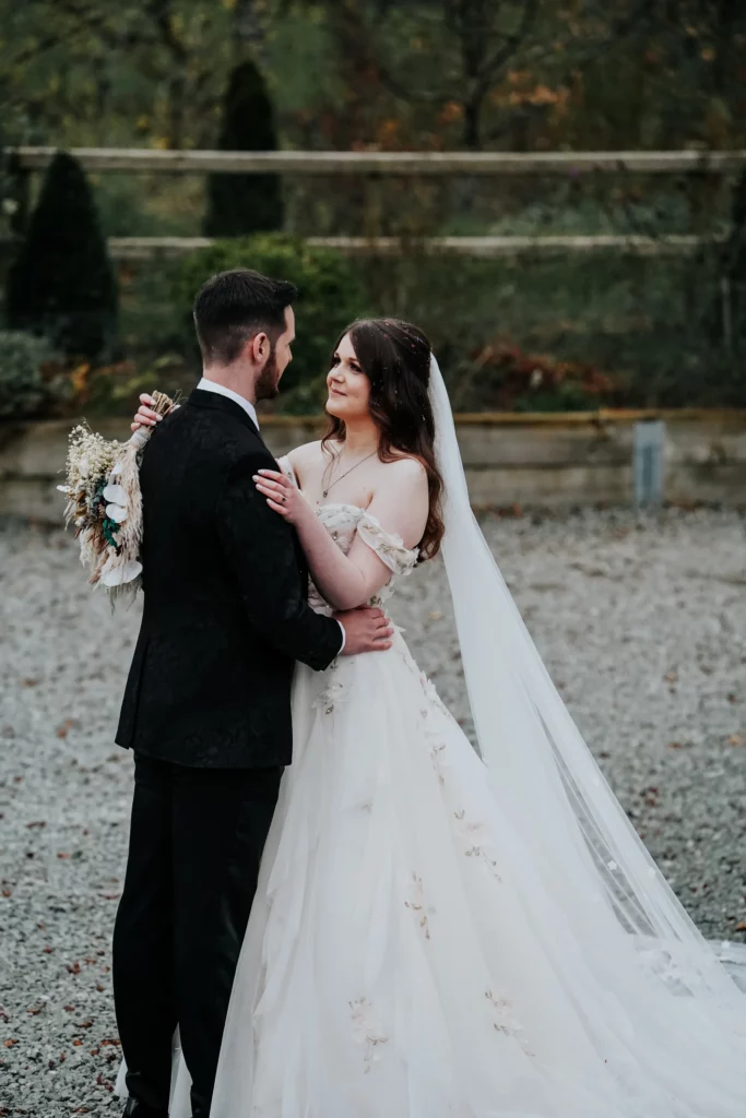A bride in a white dress and veil gazes at a groom in a black suit as they embrace outdoors on a gravel path, surrounded by greenery. The bride holds a bouquet of dried flowers. Bethany Maeve Photography