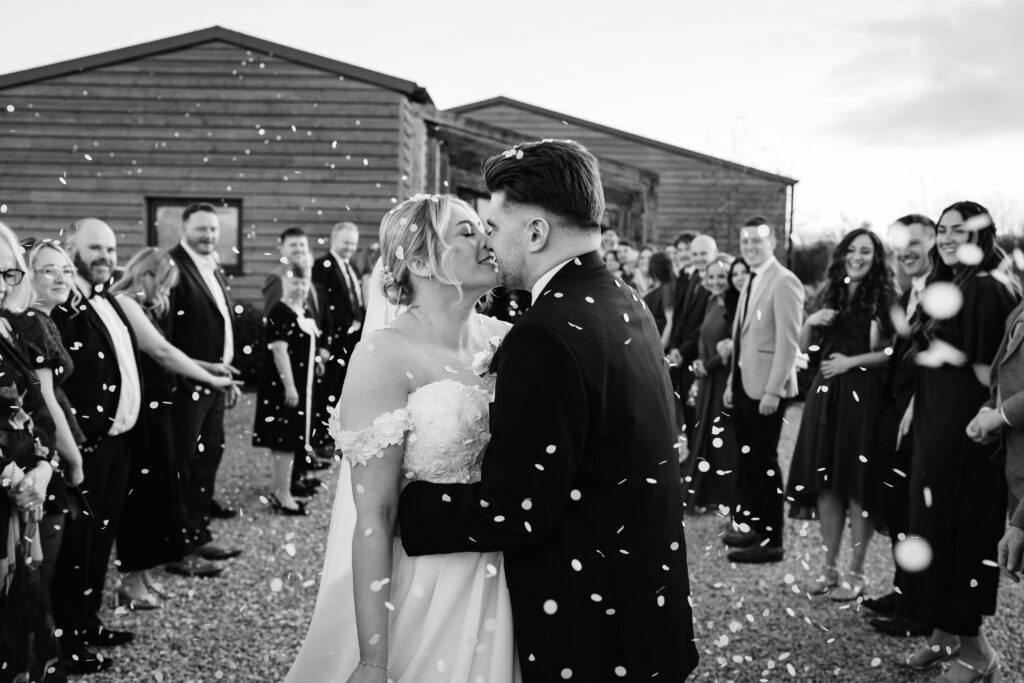 Newlyweds kissing surrounded by confetti and guests at Stretton Manor Barn for a black tie wedding